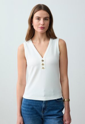 Giovane donna che indossa un top smanicato bianco a coste con tre bottoni dorati e jeans blu, in piedi contro uno sfondo chiaro.