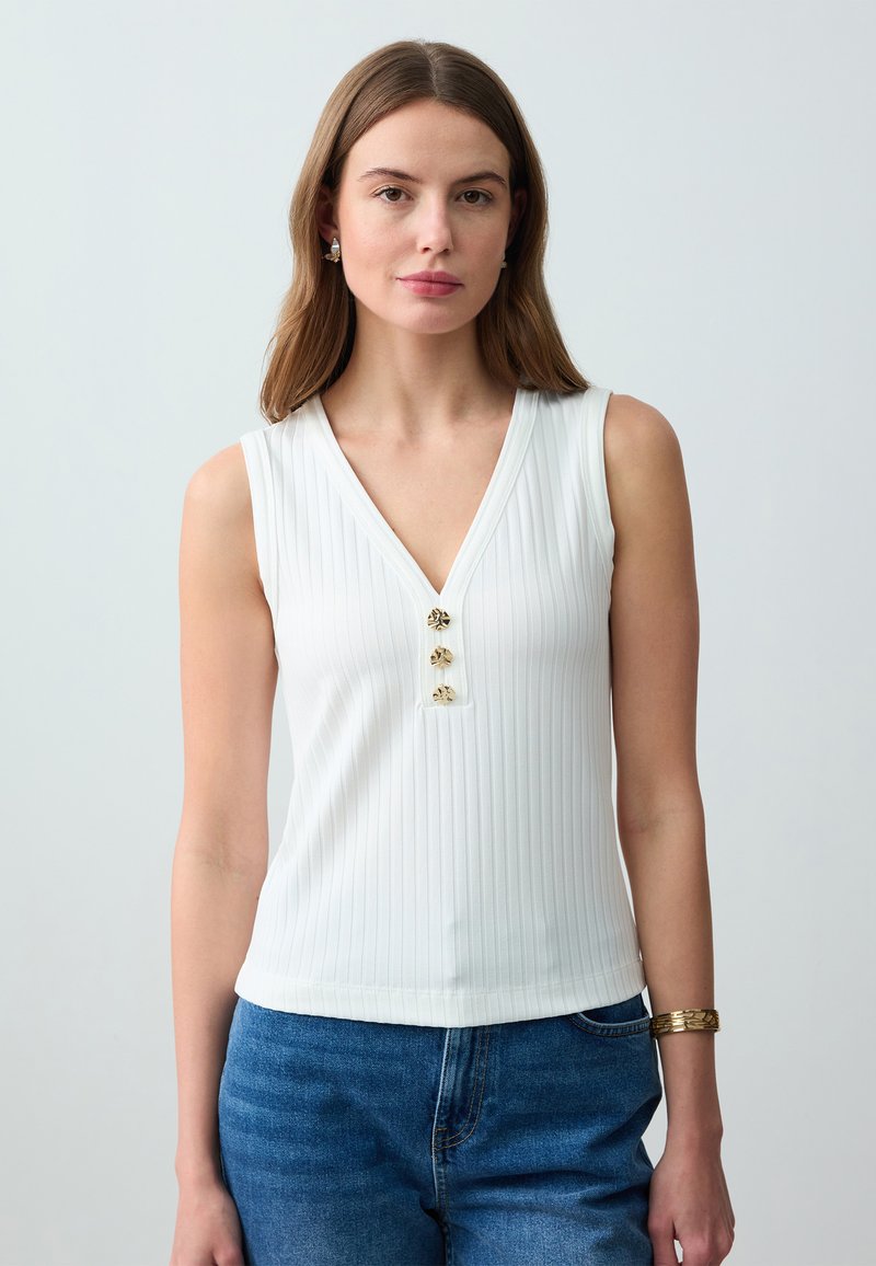 Jonge vrouw in een witte mouwloze ribbed top met drie gouden knopen en een blauwe spijkerbroek, staande tegen een effen lichte achtergrond.