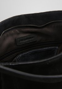 Wnętrze czarnej zamszowej torby, z tkaninowym wykończeniem, kieszenią na zamek błyskawiczny oraz metką z tłoczeniem "FRED&BRUDER". Teksturowane ciemne materiały.
