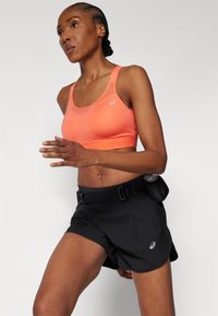 Sports-bh i lys orange med et gjennomsiktig paneldesign, kombinert med sorte løpeshorts med belte og sidelommer.