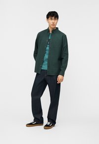 Νέος άνδρας στέκεται φορώντας σκουρό πράσινο πουκάμισο με κουμπιά πάνω από πράσινο-μπλε μπλουζάκι με γραφικά, σκούρο μπλε τζιν και μαύρα αθλητικά παπούτσια με λευκά κορδόνια και καφέ σόλες.