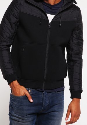 Homme portant une veste matelassée noire avec une fermeture éclair, une main dans la poche, associée à un jean bleu et une chemise foncée en dessous.