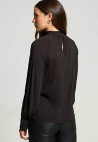 Musta pitkähihainen pusero, jossa on hienovarainen ribbikuvio, korkea kaulus nappikiinnityksellä ja pieni avokeyhole-tieto takana.