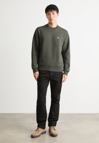 Felpa verde scuro con colletto rotondo e polsini a coste indossata su pantaloni neri. Il modello è in piedi con scarpe beige su uno sfondo neutro.
