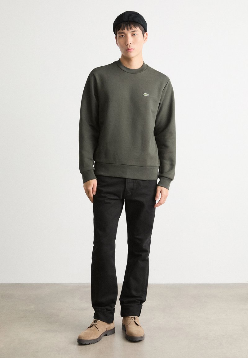 Sweatshirt vert foncé avec un col rond et des poignets côtelés porté sur un pantalon noir. Le modèle se tient debout avec des chaussures beiges sur un fond neutre.