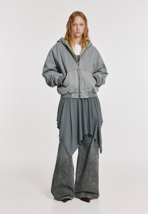 Veste à capuche grise zippée avec poignets côtelés, associée à un haut gris foncé drapé et à un jean gris à jambes larges. Texture lisse, design décontracté.