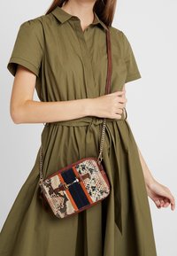 Borsa a tracolla con stampa serpente, dettagli a righe in blu navy e arancione, rifiniture marroni e una tracolla a catena, indossata con un vestito verde oliva.