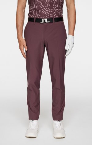 Bordeaux golfbroek met een getailleerde pasvorm, gecombineerd met een zwarte riem met een zilveren gesp. Draagt een witte handschoen en witte sneakers.