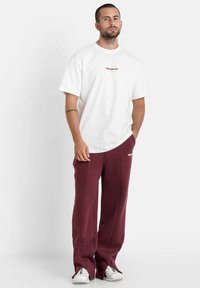 Witte t-shirt met korte mouwen en rode tekst, gecombineerd met bordeauxrode sweatpants met zijsplitten en een logo, gedragen met witte sneakers.