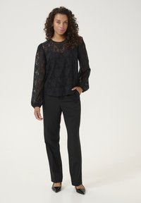 Haut en dentelle noire avec de longues manches bouffantes et un motif floral, associé à un pantalon noir ajusté et des talons noirs pointus.