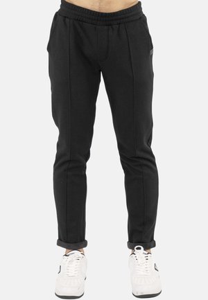 Pantalon de jogging noir slim-fit porté avec des revers roulés et des baskets blanches, montré de la taille jusqu'aux pieds sur un fond clair.