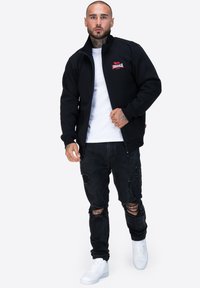 Giacca nera con zip e logo rosso, indossata sopra una t-shirt bianca; abbinata a jeans neri strappati e sneaker bianche.