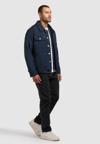 Homme marchant portant une veste bleu marine boutonnée, un t-shirt blanc, un jean noir et des baskets blanches sur un fond clair uni.