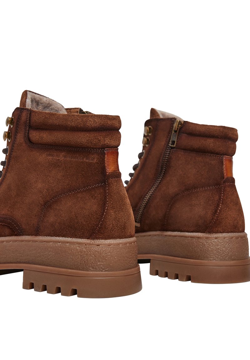 Botas de tobillo de ante marrón con una superficie texturizada, cordones en la parte delantera y cremallera lateral. Presentan una suela de goma resistente con un patrón de relieve.