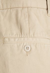 Beigefarbenes Gewebe mit glatter Textur, ausgestattet mit einer Gesäßtasche und einem runden, vierlochrigen Knopf zum Verschließen.