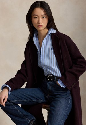 Ung kvinde iført en mørk lilla frakke, blåstribet skjorte og jeans, siddende med den ene hånd i lommen mod en neutral baggrund.