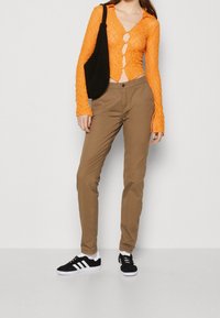 Pomarańczowa koronowa bluzka zapinana na guziki o krótkim fasonie, w połączeniu z beżowymi, dopasowanymi spodniami i czarnymi trampkami z białymi paskami, trzymająca czarną torbę.