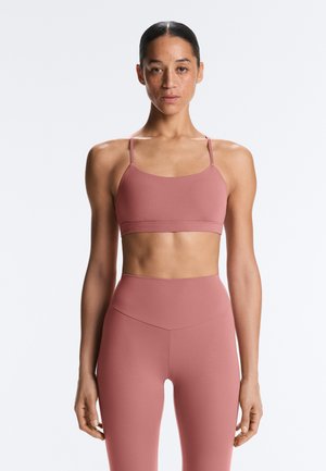 Žena s vlasmi stiahnutými dozadu, oblečená v zladenom ružovom športovom podprsenke a legínach s vysokým pásom, stojaca pred jednoliatym pozadím.