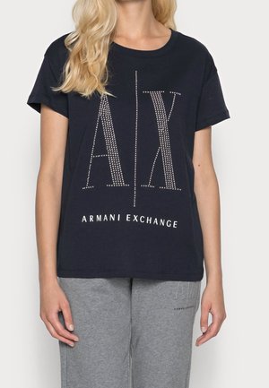 Kvinde iført en sort Armani Exchange T-shirt med stort naglet "A|X" logo og grå joggingbukser med lille logo på venstre lår.