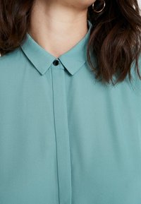 Gros plan d'une personne portant une chemise boutonnée bleu sarcelle avec un col, aux cheveux bruns mi-longs, et des boucles d'oreilles dorées en anneaux.