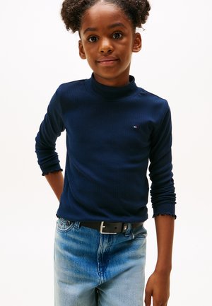 Ung pige med krøllet hår iført en mørkeblå langærmet trøje og lyseblå jeans stående mod en hvid baggrund.