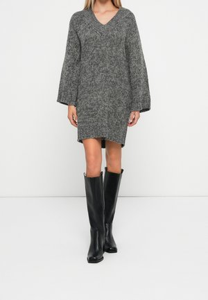 Kvinde iført en grå V-hals strikket kjole og sorte knæhøje læderstøvler, der står mod en ensfarvet hvid baggrund.