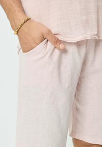 Shorts en lin rose pâle avec une coupe décontractée. Comprend des poches latérales et un tissu texturé. Une main reposant dans une poche, mettant en valeur un bracelet en or.
