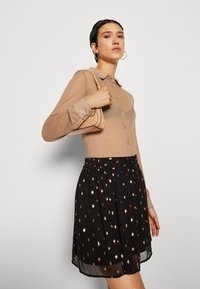 Blusa bege com botões e mangas longas combinada com uma saia preta plissada com um padrão de pontos dourados e brancos. Acessório de mala pequena.