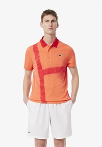 Polo arancione con colletto rosso e un design a strisce verticali rosse. Presenta una sottile texture e un logo sul lato sinistro del petto. Abbinato a pantaloni corti bianchi.