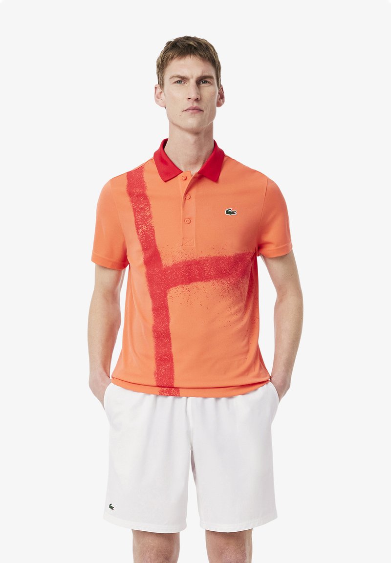 Camisa polo naranja con un collar rojo y un diseño de franjas rojas verticales. Presenta una textura sutil y un logotipo en el lado izquierdo del pecho. Combinada con pantalones cortos blancos.