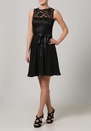 Robe noire avec un haut en dentelle, un corsage ajusté et une jupe fluide, agrémentée d'un ruban en satin à la taille. Talons noirs à brides.