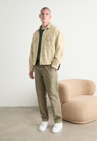 Beige Jeansjacke mit Druckknöpfen, grüner Pullover, khakifarbene Hose und weiße Sneakers. Im Hintergrund ein weich strukturierten, runden beigen Stuhl.