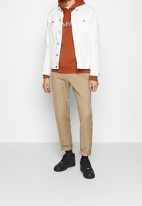 Giacca in denim bianco, felpa ruggine con logo, pantaloni beige a sigaretta e sneakers nere. Design minimalista, look casual, con colori a contrasto.