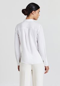 Donna con i capelli scuri raccolti in un chignon, che indossa una camicia bianca a maniche lunghe e pantaloni bianchi, rivolta di spalle su uno sfondo chiaro e semplice.