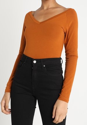 Vrouwelijk torso met een roestoranje lange mouwen V-hals top, ingestopt in hoog uitgesneden zwarte jeans met voorzakken en riemlussen.