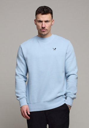 Man met kort donker haar, gekleed in een lichtblauwe trui met een kruisend peddel-logo, staand tegen een effen grijze achtergrond.