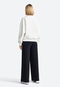 Sudadera blanca con hombros caídos, puños y dobladillo de canalé. Combinada con pantalones negros holgados y zapatillas blancas. Vista desde atrás.
