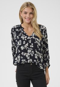 Blumenbluse in Marineblau mit weißen Blumen, leichtes Material, V-Ausschnitt und lockere Passform. Lange Ärmel mit dezenten Raffung-Details.