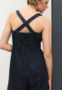 Dunkelblaues Jeans-Einteiler mit überkreuzten Trägern, tailliertem Oberteil und gerafftem Bund. Glatte Textur mit subtiler Schattierung und ohne sichtbare Verschlüsse.