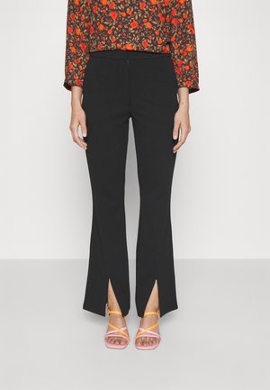 Czarne spodnie rozszerzane z przednimi rozcięciami, noszone z kwiecistym topem z długim rękawem i różowo-pomarańczowymi sandałkami na obcasie na jednolitym tle.