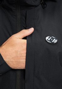 Ruka čiastočne vo vrecku na prednej strane čiernej bundy s čierno-bielym odznakom loga DC pri zipsovej priehradke na hrudi.