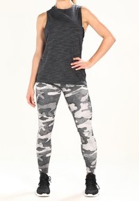 Czarny top bez rękawów o teksturowanym wykończeniu, w zestawieniu z szarymi legginsami w kamuflażowy wzór. Modelka nosi czarne buty sportowe.