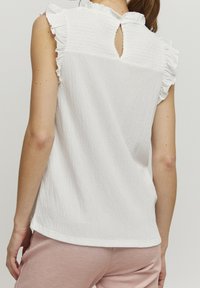 Vit topp med en texturerad tyg, volanger vid axlarna och ett nyckelhålsgrepp bak. Bärs med ljusrosa byxor.