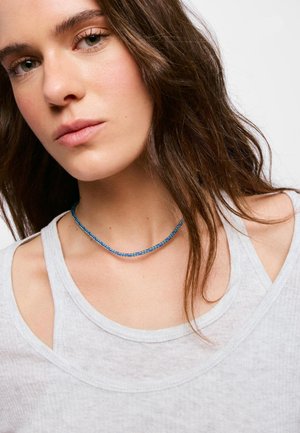 Mujer con pelo largo y castaño que lleva un top sin mangas gris claro y un collar delgado de cuentas azules, mirando ligeramente hacia un lado.