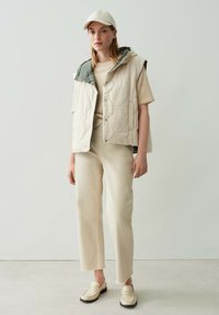 Beige geäußerte Weste mit grünem Kapuzen, beiger Pullover, hellen Hosen und cremefarbenen Loafers. Glatte Textur, minimalistisches Design und neutrale Farbpalette.