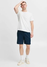 Biely bavlnený tričko v kombinácii s tmavomodrými šortkami. Model má na sebe biele ponožky a béžové tenisky s tromi pruhmi. Jednoduchý dizajn, uvoľnený strih.