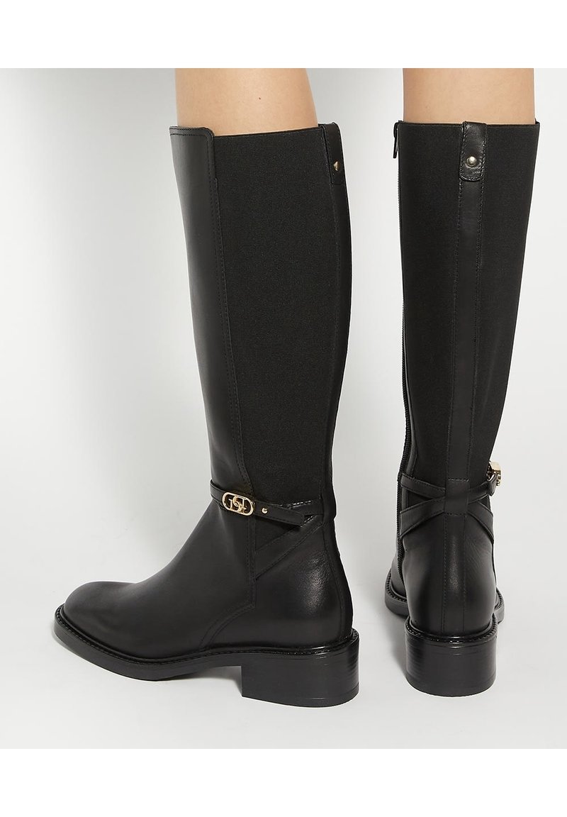 Bottes en cuir noires montantes jusqu'aux genoux avec talons bas, panneaux latéraux élastiques, et petit détail de boucle dorée sur la bride de cheville, portées sur jambes nues.