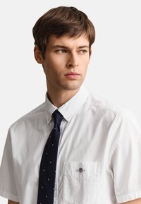 Young man with short brown hair wearing a white short-sleeve button-up shirt and dark blue tie with small white dots, looking right.