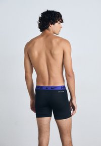 Caleçons boxers noirs avec une ceinture au logo Nike violet, présentant une coupe ajustée et une texture lisse, montrés de dos sur un modèle.