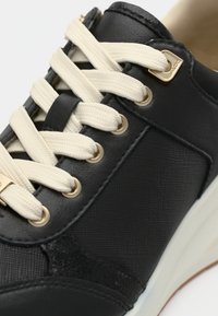 Gros plan sur une basket noire avec des lacets crème passés à travers des œillets dorés et un panneau latéral texturé.